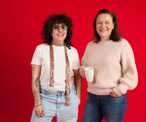 Unsere Kolleginnen lächeln in die Kamera Unsere Kolleginnen stehen vor einem roten Hintergrund und lachen in die Kamera. Sie hält eine Tasse in der Hand. Die andere trägt einen rosa Schal.