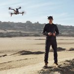 Ein Mann steht in sandiger Umgebung. Er h&auml;lt einen Drohnencontroller in der Hand und tr&auml;gt die FPV-Brille von DJI. Vor ihm fliegt die DJI FPV.
