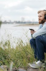 Datenvolumen Eine Person sitzt vor einem Fluss auf einem Stein und hört über das Smartphone mit Kopfhörern Musik.