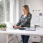 Eine Person steht an einem h&ouml;henverstellbaren Schreibtisch und arbeitet am Laptop.