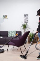Staubsauger-Vergleich Zwei Personen saugen einen Raum mit Handstaubsauger und Bodenstaubsauger, auf dem Boden steht ein Saugroboter, an einem Sofa lehnt ein Stielsauger.