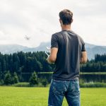 Eine Person steht auf einer Wiese und l&auml;sst eine Drohne steigen, die am Horizont verschwommen &uuml;ber einer Baumreihe fliegt.