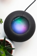 HomePod Spotify Ein schwarzer Apple HomePod steht neben zwei Pflanzen auf einem weißen Tisch.
