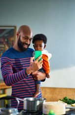 Eine Person mit einem Kind auf dem Arm steht vor einer Kücheninsel. In der freien Hand hält sie ein Smartphone, in das die beiden lächelnd hineinschauen. Eine Person mit einem Kind auf dem Arm steht vor einer Kücheninsel. In der freien Hand hält sie ein Smartphone, in das die beiden lächelnd hineinschauen.