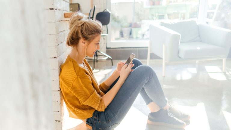 Eine Person lehnt sitzend an einer Wand in einem Wohnzimmer und schaut auf das Smartphone in ihren H&auml;nden.