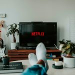 Blick auf einen Fernseher, auf dem Netflix l&auml;uft. Im Vordergrund sind die F&uuml;&szlig;e einer Person auf einem Tisch zu sehen.