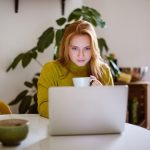 Eine Person mit einer Kaffeetasse in der Hand sitzt vor einem Laptop.