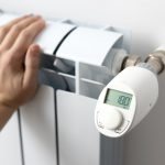 Eine Person fasst an eine Heizung, an der ein elektronisches Thermostat sitzt.