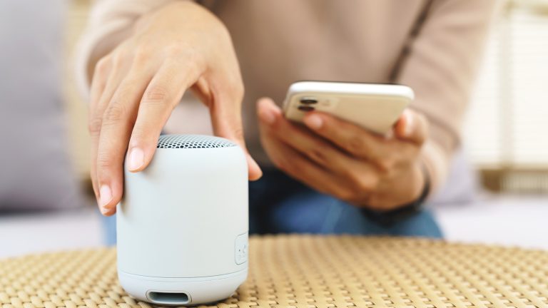 Eine Person hat ein Smartphone in der Hand. Mit der anderen Hand stellt sie einen kleinen Wireless Speaker auf einen Korbtisch.