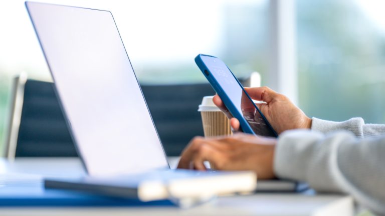 Eine Person h&auml;lt ein iPhone in der Hand und scheint an einem Schreibtisch zu sitzen.