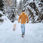 Eine Person l&auml;uft auf einem schneebedeckten Waldweg mit einem Hund spazieren und ist dabei winterlich gekleidet.