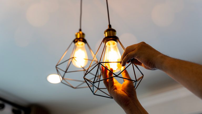 Eine Person schraubt eine Gl&uuml;hbirne in eine, an der Decke h&auml;ngende, Lampe.