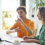 Zwei Personen sitzen an einem Tisch und schauen gemeinsam in einen Laptop. Eine der beiden Personen kommuniziert w&auml;hrenddessen mit ihren H&auml;nden in Geb&auml;rdensprache.
