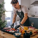 Eine Person in schwarzer Sch&uuml;rze und Handschuhen bereitet auf einem Elektrogrill Gem&uuml;se und Fleisch zu. Neben ihm steht eine Person mit einem Glas Wei&szlig;wein. Im Hintergrund eine helle, minimalistisch eingerichtete K&uuml;che mit Dunstabzugshaube.