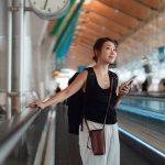 Eine Person steht auf einem Laufband im Flughafen. Mit der einen Hand h&auml;lt sie sich fest, in der anderen h&auml;lt sie ein Smartphone.