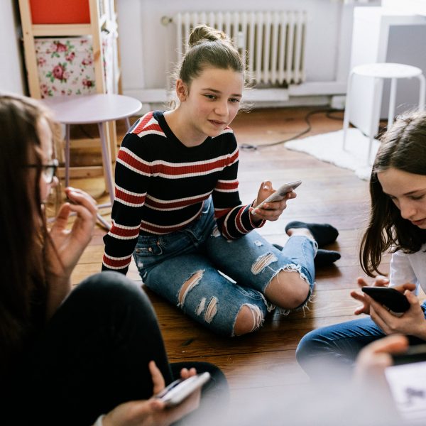 Drei junge Personen sitzen auf dem Boden in einem Zimmer. Alle drei halten ein Smartphone in der Hand.