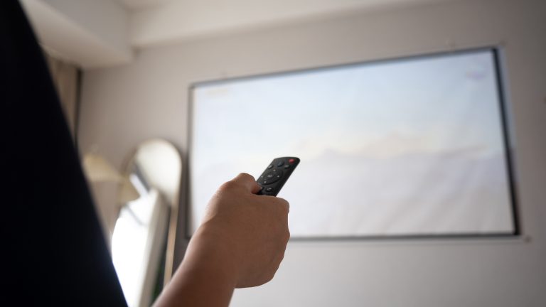 Es ist von hinten der Arm einer Person zu sehen, die eine Fernbedienung auf einen Beamer richtet.